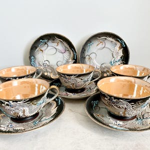 May include: A set of six teacups and saucers with a black and white dragon design. The cups and saucers have a gold trim and are decorated with blue and white accents.