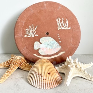 May include: A terracotta round plate with a painted fish and seaweed design. The plate is surrounded by three seashells, including a starfish and a scallop shell.