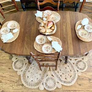 May include: A wooden dining table set for a meal, featuring woven placemats, floral patterned plates, white napkins, and a basket of bread. The table is surrounded by wooden chairs and a decorative jute rug.