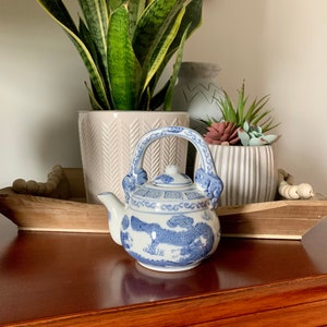 May include: A small blue and white porcelain teapot with a dragon design. The teapot has a handle and spout and is sitting on a wooden surface.