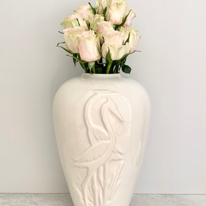 May include: White ceramic vase with a raised design of two birds. The vase is filled with a bouquet of pale pink roses.