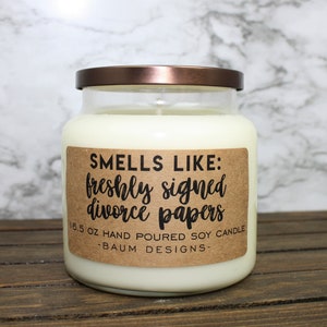 May include: A clear glass jar candle with a copper lid and a brown paper label that reads "SMELLS LIKE: freshly signed divorce papers 6.5 OZ HAND POURED SOY CANDLE -BAUM DESIGNS-"
