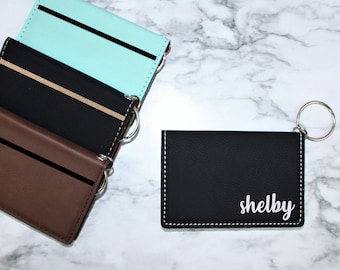 May include: Four leather card holders with a key ring. The card holders are in brown, black, and turquoise. One card holder has the name "shelby" written on it in white.