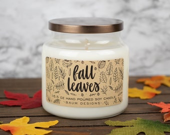 May include: A white glass jar candle with a brown label that says "fall leaves" and "16.5 oz hand poured soy candle -BAUM DESIGNS-." The candle is sitting on a wooden surface with fall leaves scattered around it.