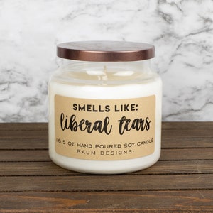 May include: A white soy candle in a clear glass jar with a copper lid. The label reads "Smells Like: liberal tears" and "16.5 oz hand poured soy candle -BAUM DESIGNS-."