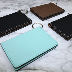 May include: Four leather keychains in different colors: black, brown, turquoise, and black. Each keychain has a metal ring and a stitched edge.