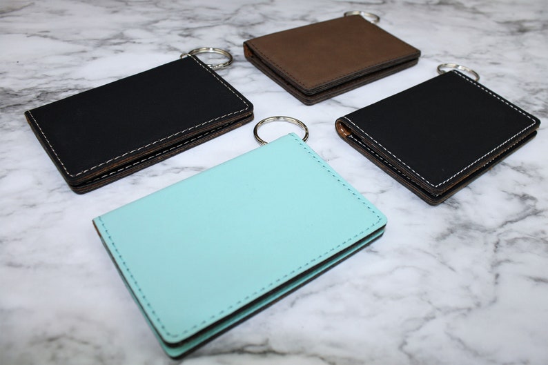 May include: Four leather keychains in different colors: black, brown, turquoise, and black. Each keychain has a metal ring and a stitched edge.