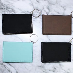 May include: Four keychains with different colored leather card holders. The keychains are silver and the card holders are black, brown, light blue, and black.