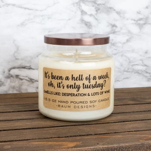 May include: A clear glass jar candle with a copper lid. The label reads "It's been a hell of a week, oh, it's only tuesday?" and "Smells like: Desperation & Lots of Wine". The candle is 16.5 ounces and hand-poured soy.