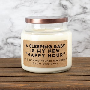 K&ouml;nnte beinhalten: Eine wei&szlig;e Sojawachskerze in einem durchsichtigen Glasgef&auml;&szlig; mit einem kupferfarbenen Deckel. Das Etikett lautet "A sleeping baby is my new "Happy Hour"" 16.5 oz hand poured soy candle -Baum Designs-.