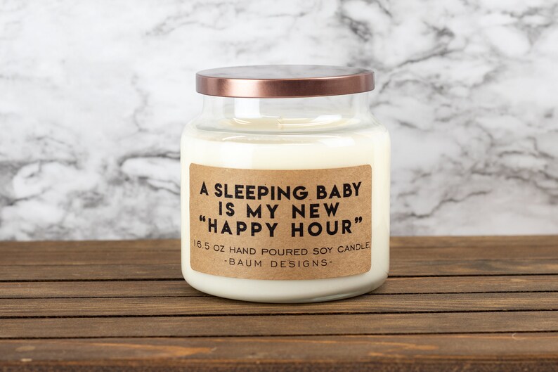 K&ouml;nnte beinhalten: Eine wei&szlig;e Sojawachskerze in einem durchsichtigen Glasgef&auml;&szlig; mit einem kupferfarbenen Deckel. Das Etikett lautet "A sleeping baby is my new "Happy Hour"" 16.5 oz hand poured soy candle -Baum Designs-.