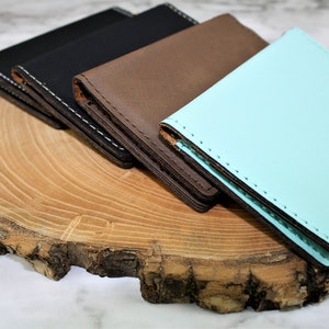 May include: Four leather wallets stacked on a wooden surface. The wallets are in different colors: black, brown, and light blue.