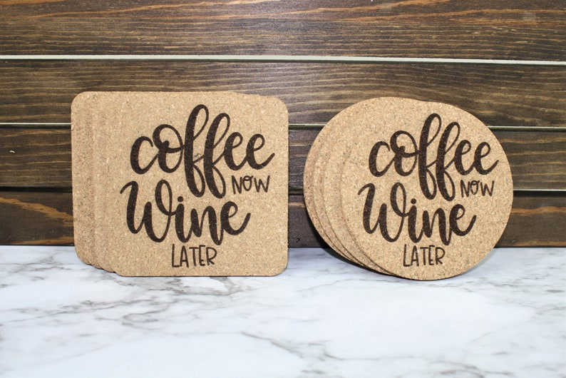 May include: Set of six round cork coasters with the text "coffee now wine later" in black script font. The coasters are stacked on a white marble surface.