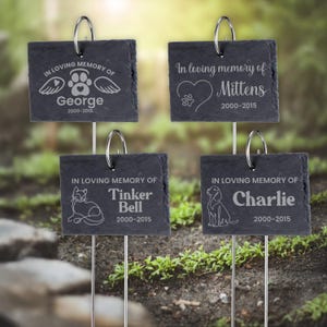 May include: Four black slate pet memorial garden stakes. Each stake has a silver metal loop and reads "In loving memory of" followed by a pet's name and dates. The stakes feature a paw print, cat, dog, and angel wings.