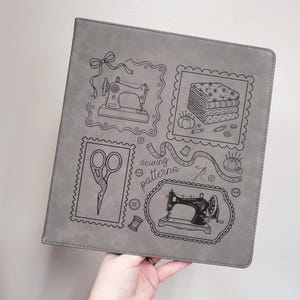 May include: A square gray binder featuring black line art illustrations of sewing supplies, including two sewing machines, a stack of fabric, scissors, a pincushion, and a measuring tape, with the text sewing patterns printed in the center.