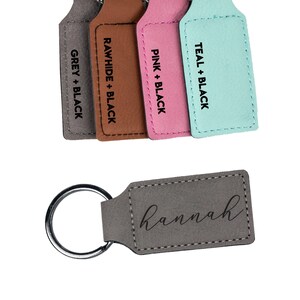May include: A gray leather keychain with a silver ring and the name "hannah" engraved on it. The keychain is shown next to four other keychains in different colors: gray, brown, pink, and teal.