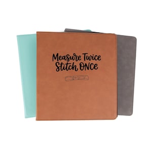 May include: Three rectangular binders in light blue, tan, and gray. The front tan binder features the black text "Measure Twice Stitch ONCE" above a small graphic of a measuring tape with the numbers one, two, and three clearly visible.