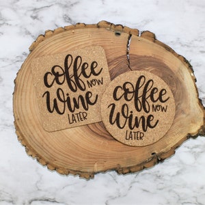 May include: Two cork coasters with the text "coffee now wine later" in black script. The coasters are on a wooden slice.