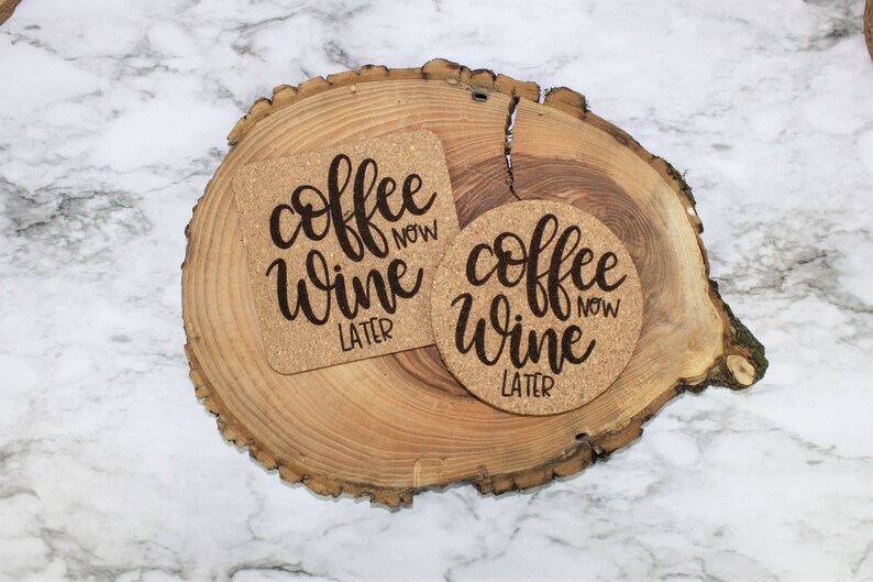 May include: Two cork coasters with the text "coffee now wine later" in black script. The coasters are on a wooden slice.