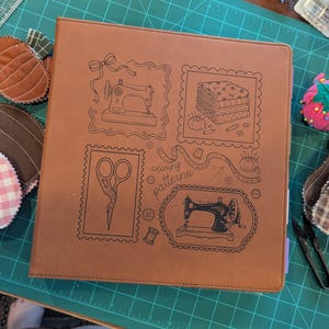 May include: A tan faux leather binder featuring black line art illustrations of sewing machines, scissors, a stack of fabric, and a measuring tape. The cover includes the text "sewing patterns" in a cursive font, surrounded by decorative frames.