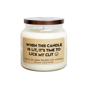 May include: A white soy candle in a clear glass jar with a brown lid. The label is brown with the text "WHEN THIS CANDLE IS LIT, IT'S TIME TO LICK MY CLIT" and a tongue sticking out emoji. The label also says "16.5 OZ HAND POURED SOY CANDLE -BAUM DESIGNS-"