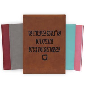 May include: A collection of journals in various colors, including red, gray, brown, teal, and pink. The central journal is brown with the text "Shezby's June Journal" and a heart symbol. The journals are arranged side by side.