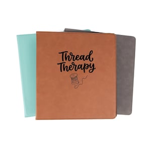 May include: Three square binders with smooth covers in light blue, tan, and gray. The front tan binder features the black text Thread Therapy in a script font above a line drawing of a thread spool with a loose strand of thread trailing below.