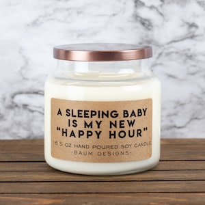 K&ouml;nnte beinhalten: Eine Glas-Kerze in einem Glas mit einem braunen Etikett, auf dem "A Sleeping Baby Is My New "Happy Hour"" und "16.5 oz Hand Poured Soy Candle - Baum Designs -" steht.