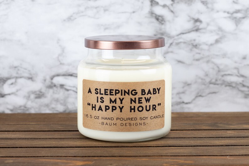 K&ouml;nnte beinhalten: Eine Glas-Kerze in einem Glas mit einem braunen Etikett, auf dem "A Sleeping Baby Is My New "Happy Hour"" und "16.5 oz Hand Poured Soy Candle - Baum Designs -" steht.