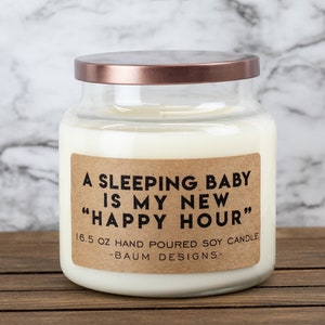 K&ouml;nnte beinhalten: Eine Kerze in einem Glasgef&auml;&szlig; mit einem braunen Etikett, auf dem "A sleeping baby is my new "happy hour"" steht. Das Etikett tr&auml;gt au&szlig;erdem den Text "16.5 oz hand poured soy candle -Baum Designs-".