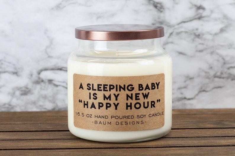 K&ouml;nnte beinhalten: Eine Kerze in einem Glasgef&auml;&szlig; mit einem braunen Etikett, auf dem "A sleeping baby is my new "happy hour"" steht. Das Etikett tr&auml;gt au&szlig;erdem den Text "16.5 oz hand poured soy candle -Baum Designs-".