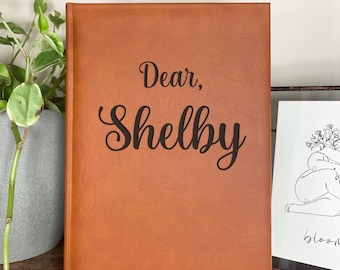 May include: A brown leather-bound journal with the text "Dear, Shelby letters to my daughter" engraved on the front cover.