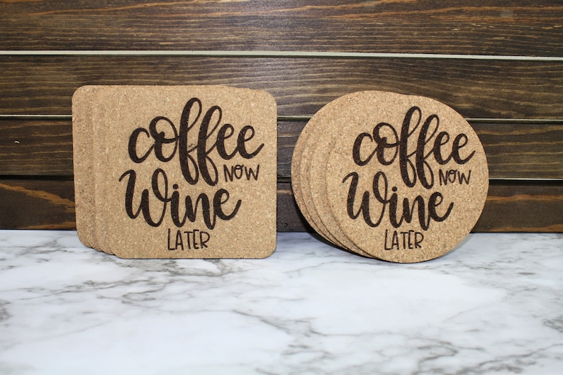 May include: Two sets of cork coasters with the text "coffee now wine later" in a cursive font. The coasters are on a white marble surface.