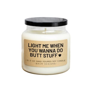 May include: A glass jar candle with a black lid and a brown paper label that reads "LIGHT ME WHEN YOU WANNA DO BUTT STUFF ♥" and "16.5 OZ HAND POURED SOY CANDLE -BAUM DESIGNS-"