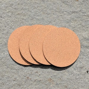 May include: Four round cork coasters in a light brown color. The coasters are stacked on top of each other.