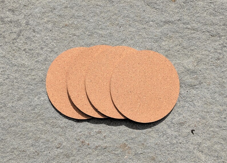May include: Four round cork coasters in a light brown color. The coasters are stacked on top of each other.