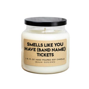 May include: A clear glass jar candle with a black lid. The label is brown with black text that reads "Smells Like You Have (Band Name) Tickets". The label also includes the text "16.5 oz Hand Poured Soy Candle - Baum Designs -."