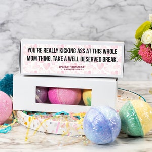 May include: A white box set containing three bath bombs, with a second box on top that reads "YOU'RE REALLY KICKING ASS AT THIS WHOLE MOM THING, TAKE A WELL DESERVED BREAK." The bath bombs are pink, purple, and yellow. A bouquet of flowers is in the background.