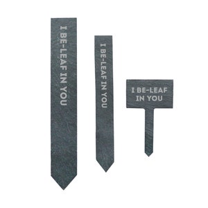 May include: Three slate garden markers with the text "I Be-Leaf In You" in white lettering. The markers are different sizes and shapes, with two being long and narrow and one being a small rectangular sign with a stake.