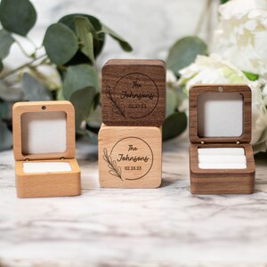 May include: Three wooden ring boxes with a hinged lid. The boxes are engraved with the text "The Johnsons" and the date "02.23.23". One box is light wood, one is dark wood, and one is a stack of two boxes, one light and one dark.