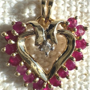 May include: A heart-shaped pendant crafted from polished yellow gold, featuring a border of round, deep pink rubies set in prongs. A single, small, clear round-cut diamond is centered at the top of the heart, suspended from a gold bail.