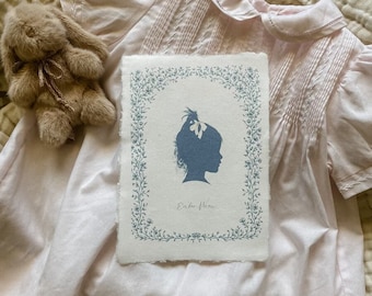 Può includere: Un vestitino bianco per neonata con un bordo floreale blu intorno a una silhouette di una bambina con un fiocco nei capelli. Il testo "Ein für immer" è scritto sotto la silhouette.