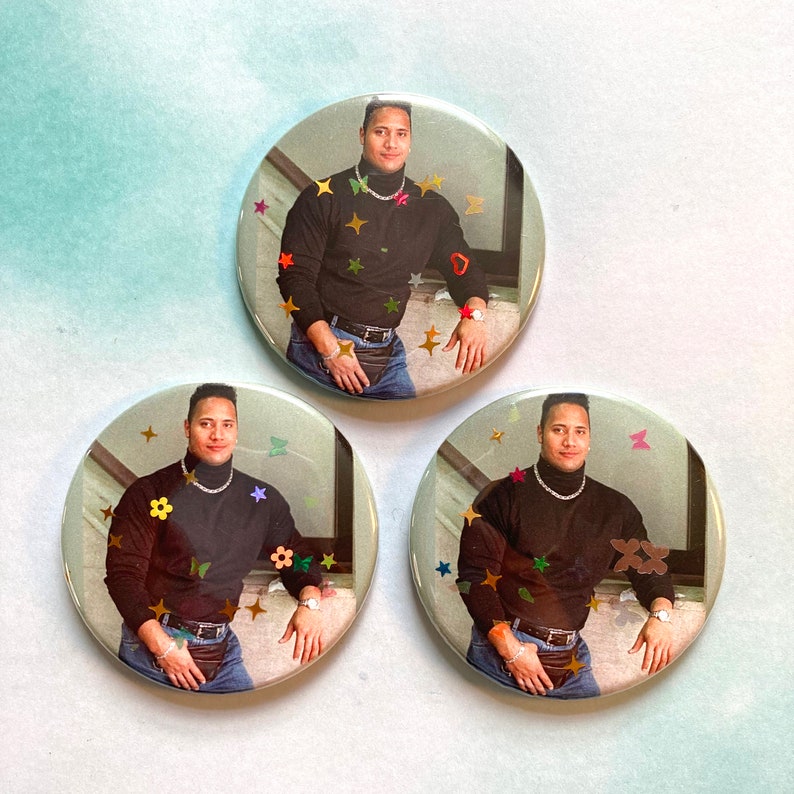 May include: Three round buttons with a black and white image of a man wearing a black turtleneck shirt and blue jeans. The buttons are decorated with colorful glitter and stars.