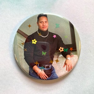 May include: A black mock neck shirt with a gold chain necklace and blue jeans. The shirt has a variety of colorful flowers and butterflies on it.