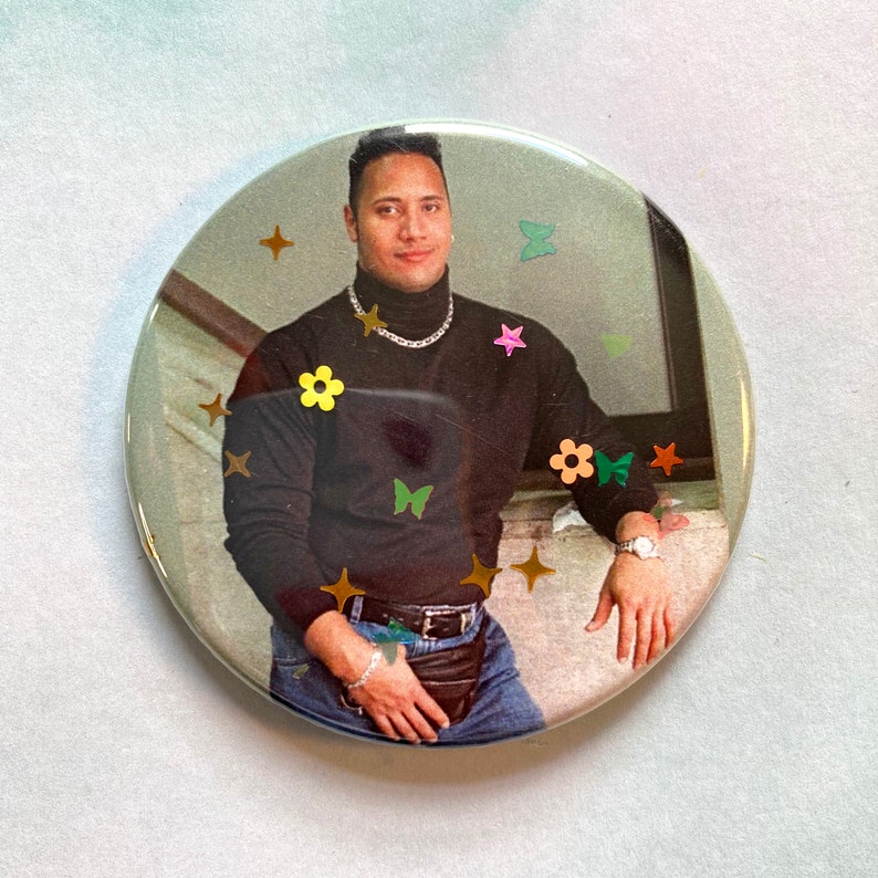 May include: A black mock neck shirt with a gold chain necklace and blue jeans. The shirt has a variety of colorful flowers and butterflies on it.