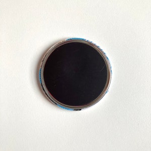 May include: The back of a round button with a black magnetic surface and a blue and white patterned border.