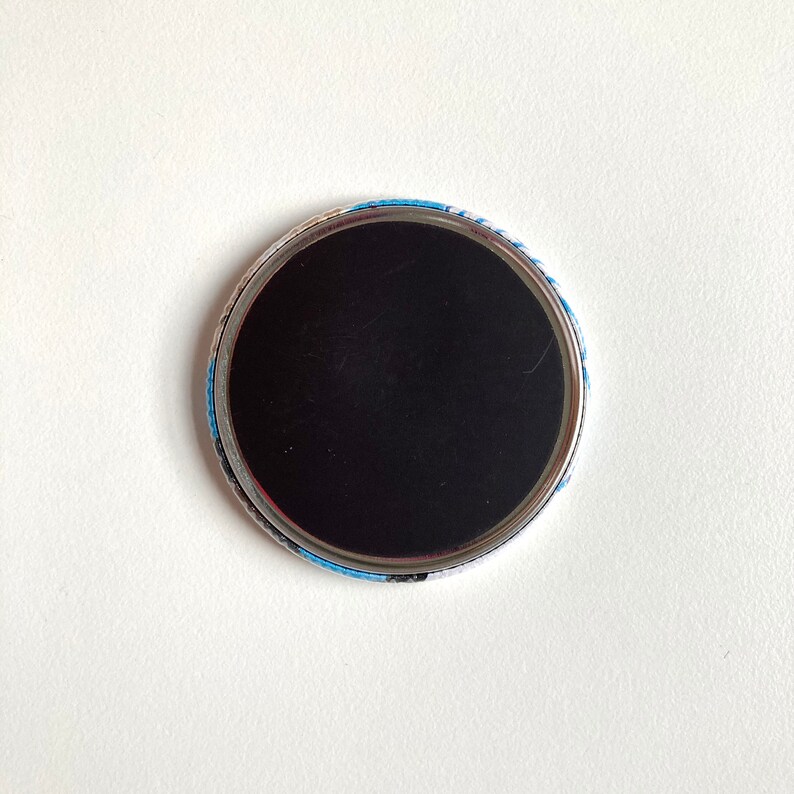 May include: The back of a round button with a black magnetic surface and a blue and white patterned border.