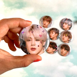 Op de afbeelding: Een set van acht ronde knoopsluitingen met verschillende foto's van een K-pop idool. De knoopsluitingen zijn versierd met glitter en kleine hartjes.