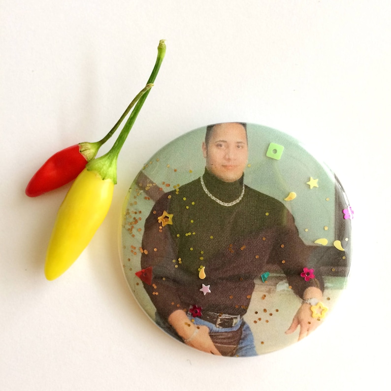 May include: A round button with a photo of a man wearing a black turtleneck sweater and blue jeans. The button is decorated with gold glitter and colorful sequins.