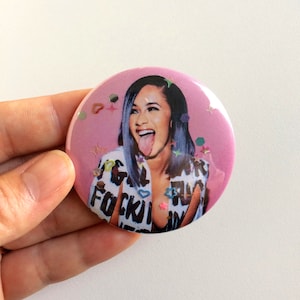May include: A pink button with a picture of a woman with blue hair sticking her tongue out. The background is pink with colorful glitter.